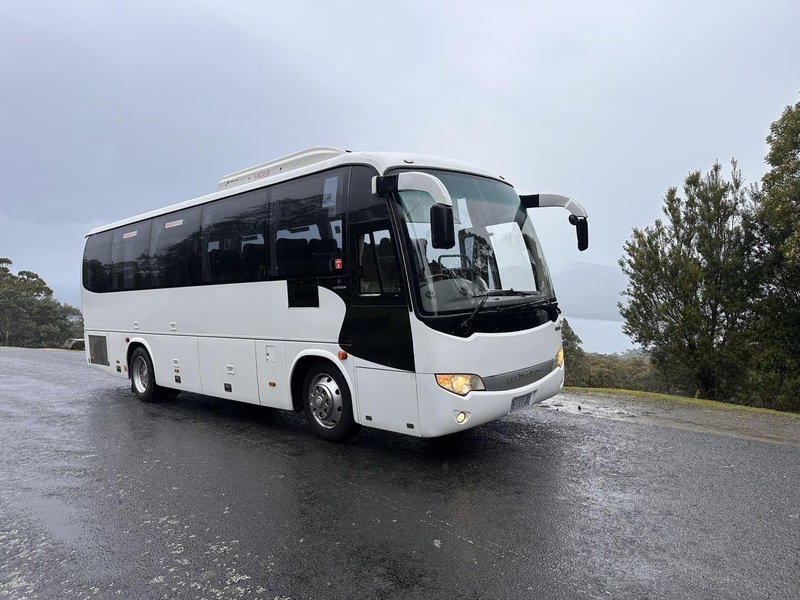 White coach with its headlights on, parked on a wet road under grey skies
