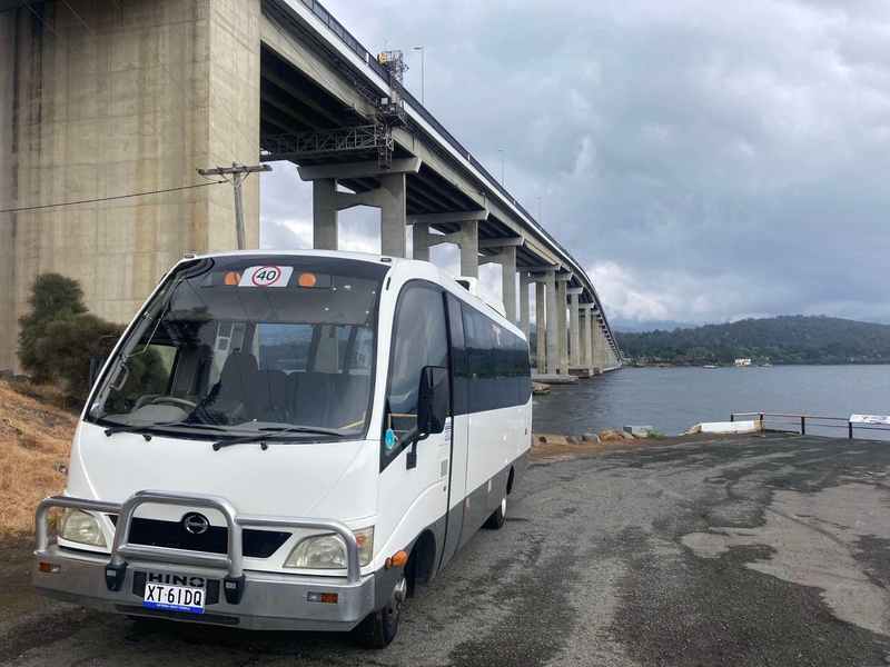 White minibus with a bull bar parked beneath a tall concrete bridge beside the water