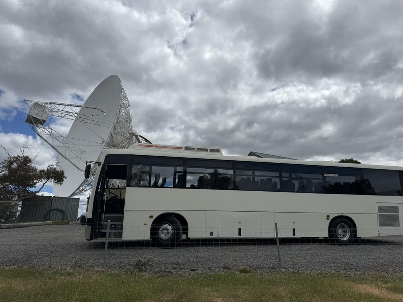 Side view of a white coach beside a large satellite dish under cloudy skies