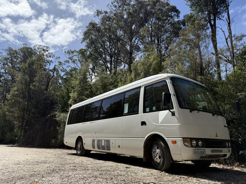 White minibus parked on gravel in front of tall gum trees and blue sky