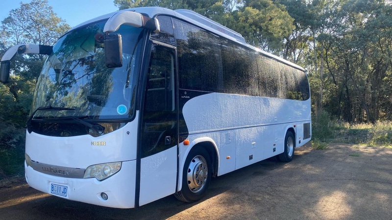 Front three-quarter view of a white coach parked on a dirt track among bush