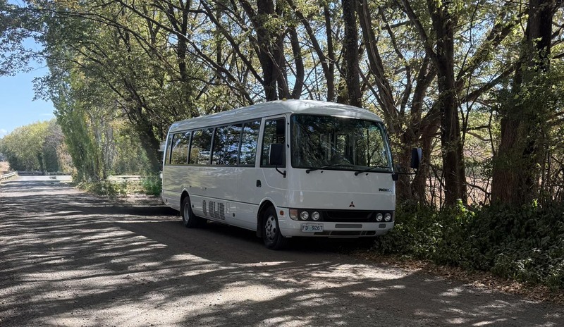 White minibus parked in dappled shade beside trees on a gravel road