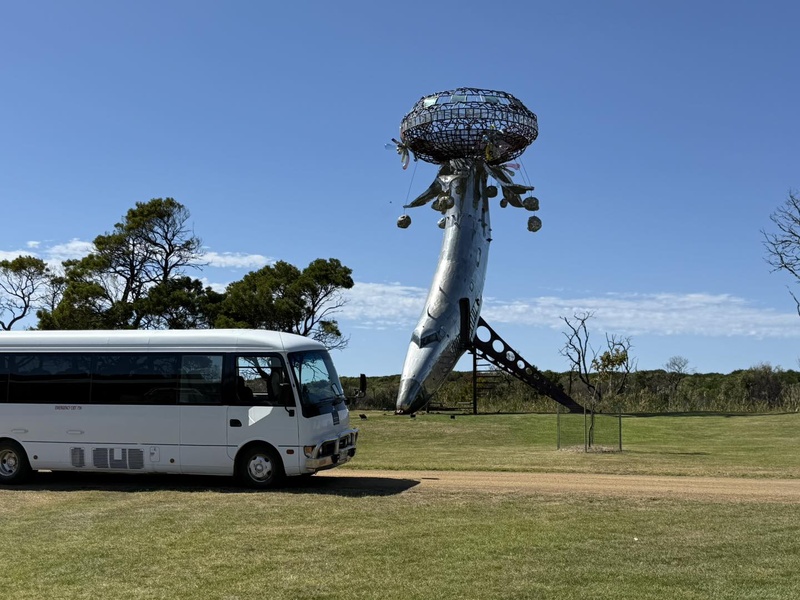 White minibus parked on a grassy field beside a tall metal sculpture