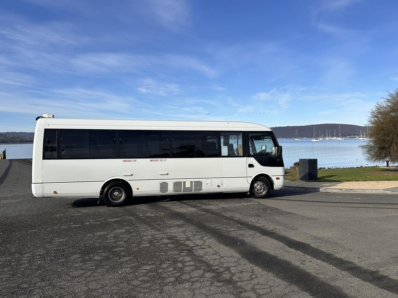 White minibus parked side-on at the waterfront, with boats and hills across the bay